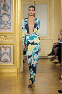 Model walking a runway in a long navy floral jumpsuit with a deep V neckline in a gilded, ornate room.