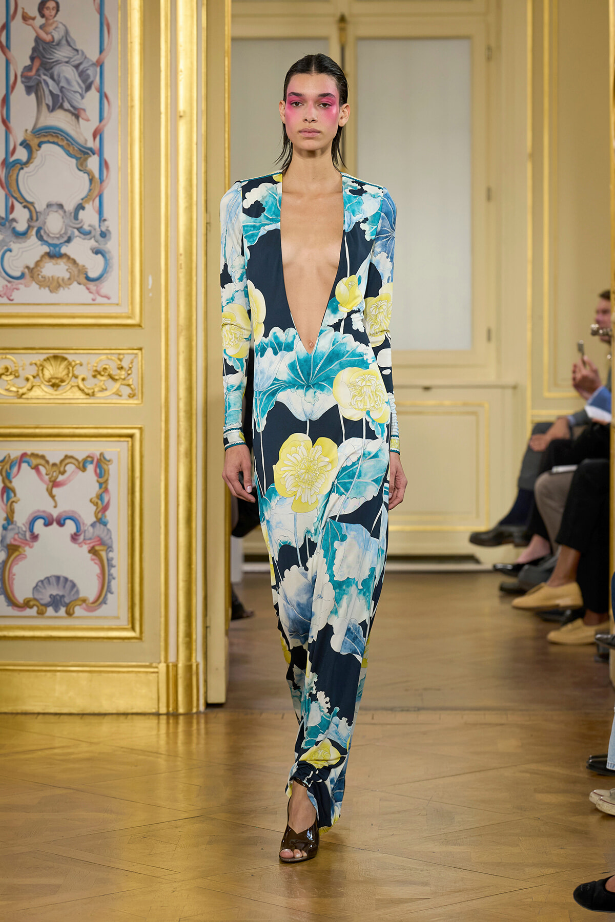 Model walking a runway in a long navy floral jumpsuit with a deep V neckline in a gilded, ornate room.