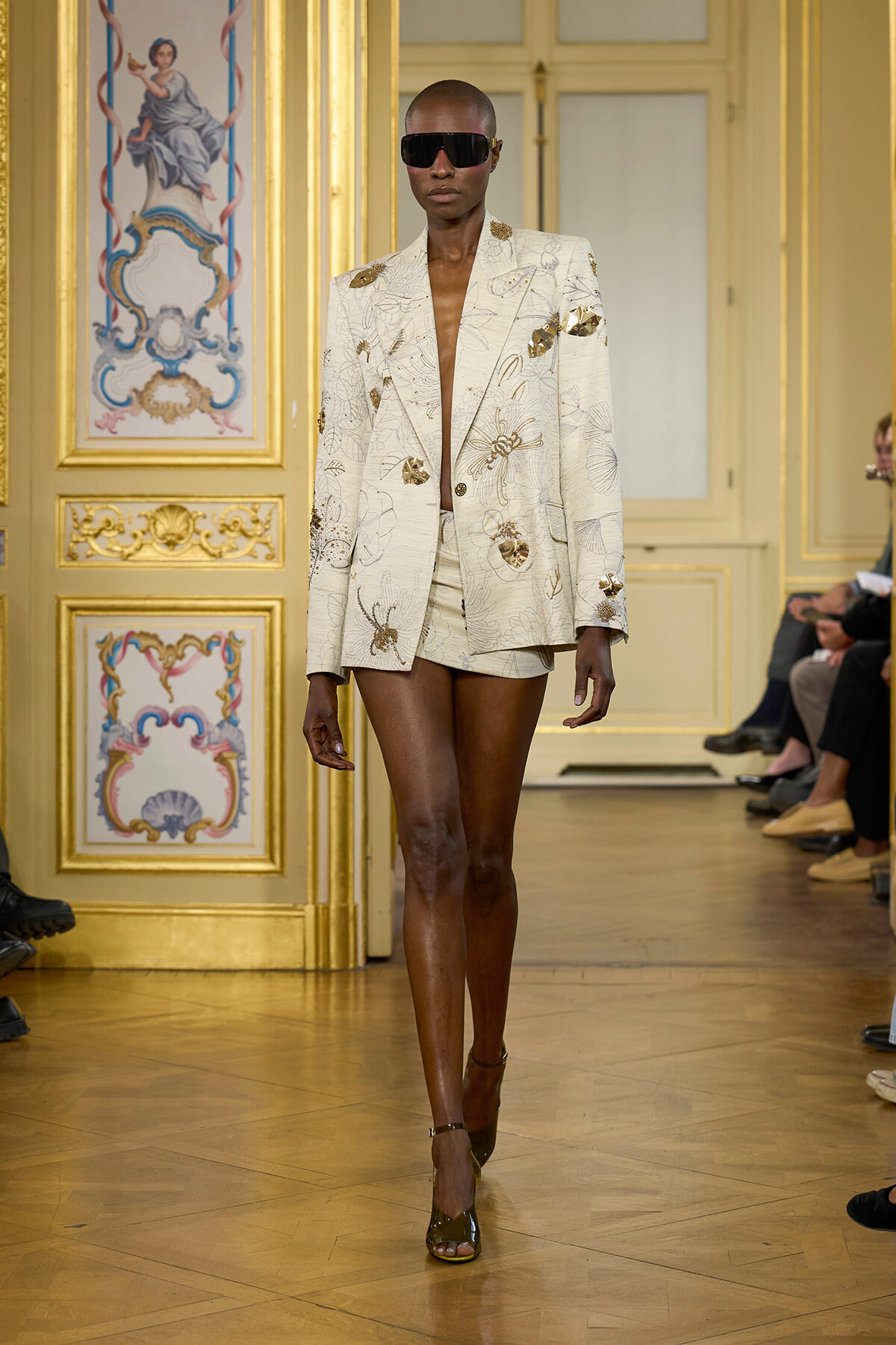 Model struts a cream floral-embroidered blazer with matching shorts on a gold-decorated runway. She wears oversized sunglasses and heels.