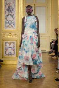 Model walks the runway in a pastel floral ballgown with a high-low, voluminous skirt in a gilded, ornate room.