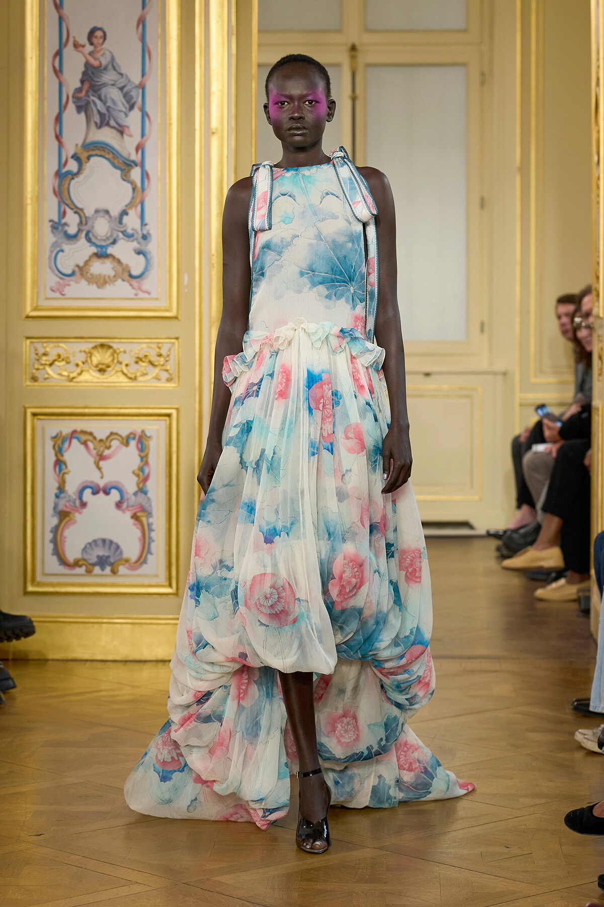 Model walks the runway in a pastel floral ballgown with a high-low, voluminous skirt in a gilded, ornate room.