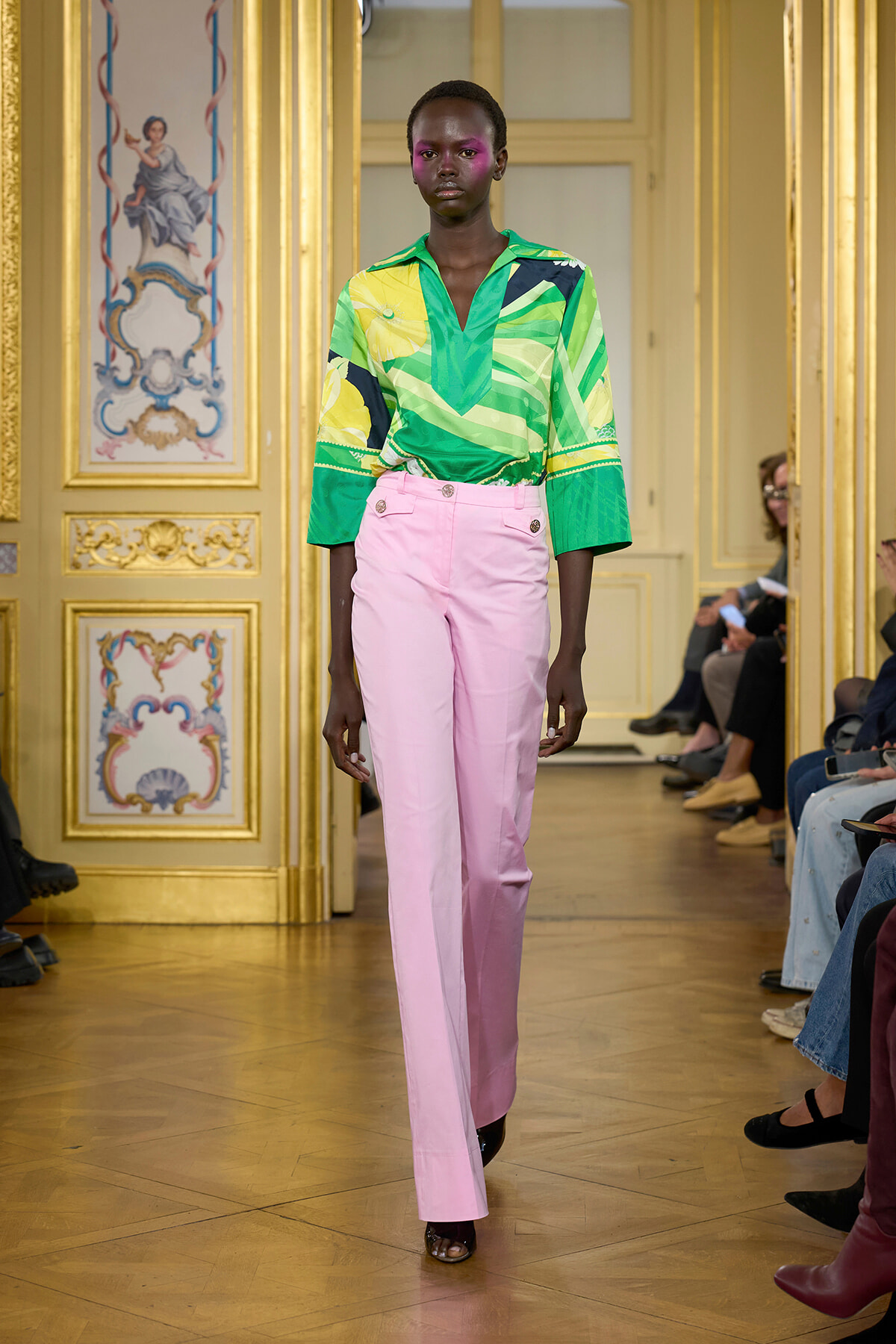 Model walks a runway in a green geometric blouse and light pink high-waisted pants in a gilded, ornate room.