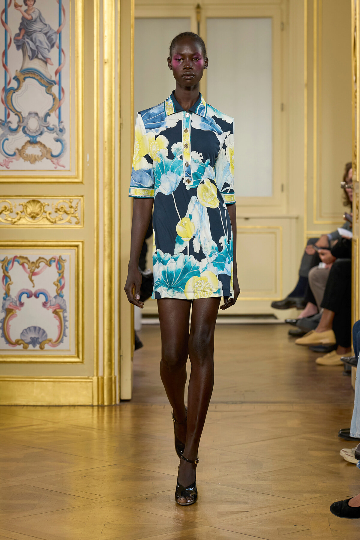 Model walking a fashion show in a navy floral dress with yellow flowers, short sleeves, and legs visible, in a gilded ornate room with gold trim and decorative panels.