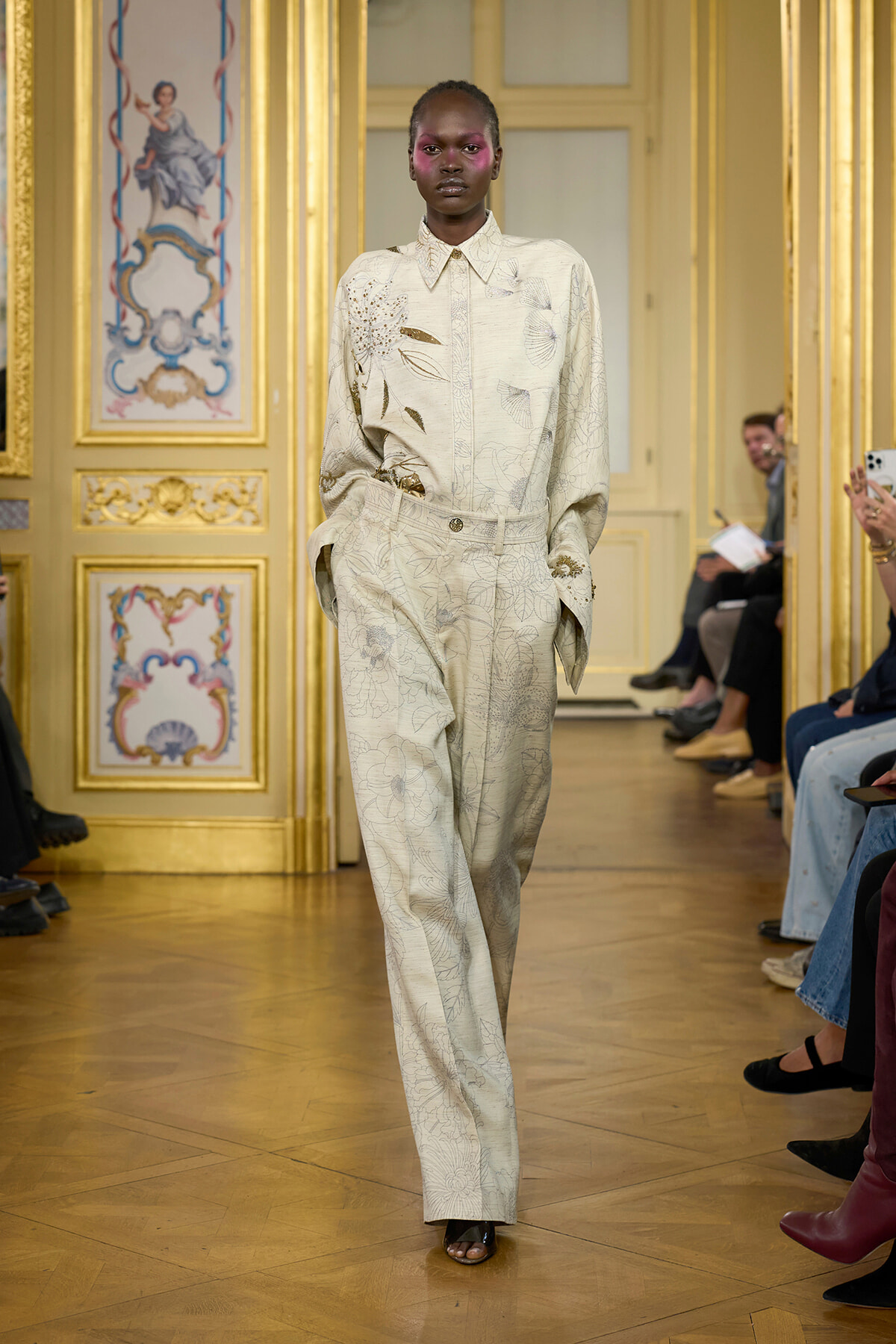 Model on a runway wearing a pale cream floral suit with a buttoned shirt and wide-leg trousers, walking through a gilded, ornate interior.