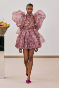 Model walking a runway in a pink floral dress with oversized puff sleeves and pink rose-adorned sandals.