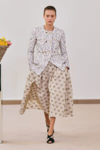 Model wearing a white speckled tweed jacket with gold buttons and a beige floral midi skirt, standing indoors with a fruit basket on the left.