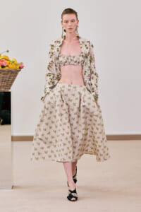 Model on a runway wearing a cream floral two-piece: ruffled bandeau top, voluminous knee-length skirt with butterfly print, and a matching embroidered jacket; a basket of lemons sits to the left.