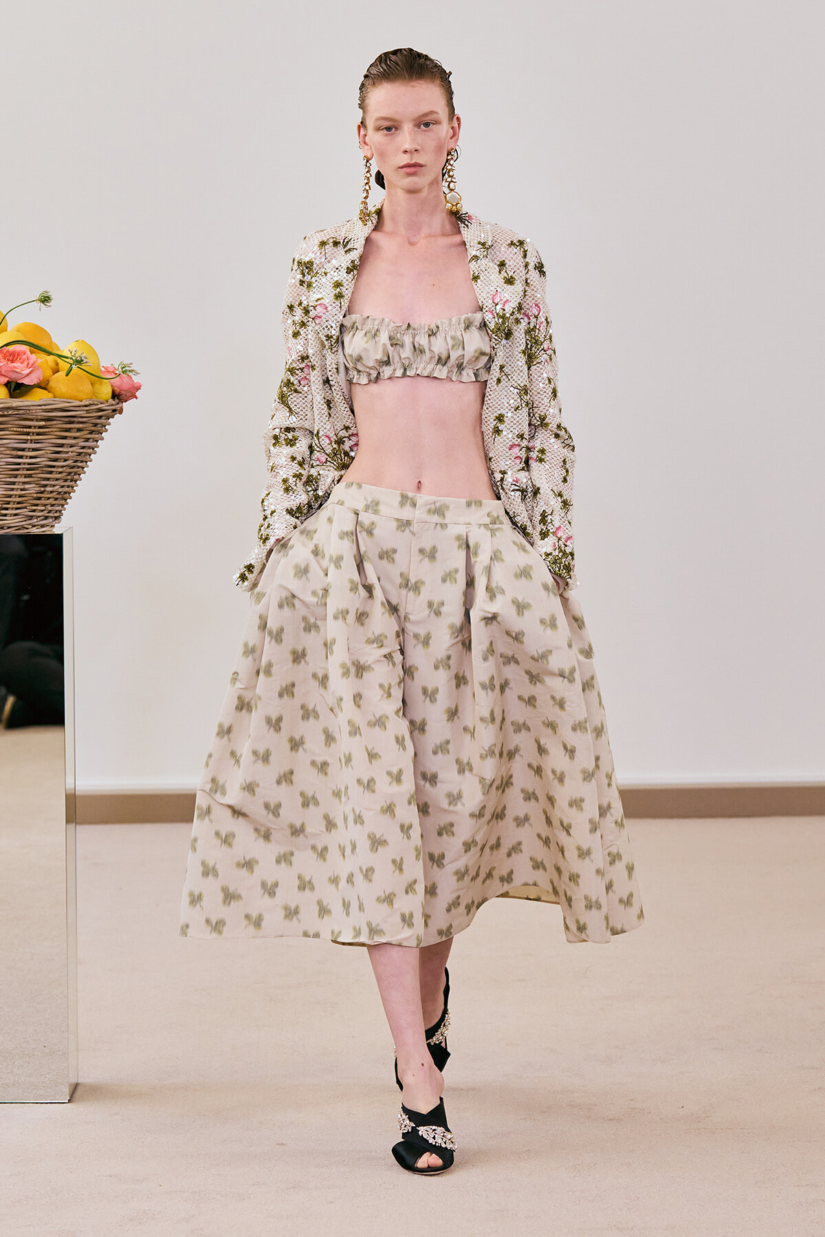 Model on a runway wearing a cream floral two-piece: ruffled bandeau top, voluminous knee-length skirt with butterfly print, and a matching embroidered jacket; a basket of lemons sits to the left.