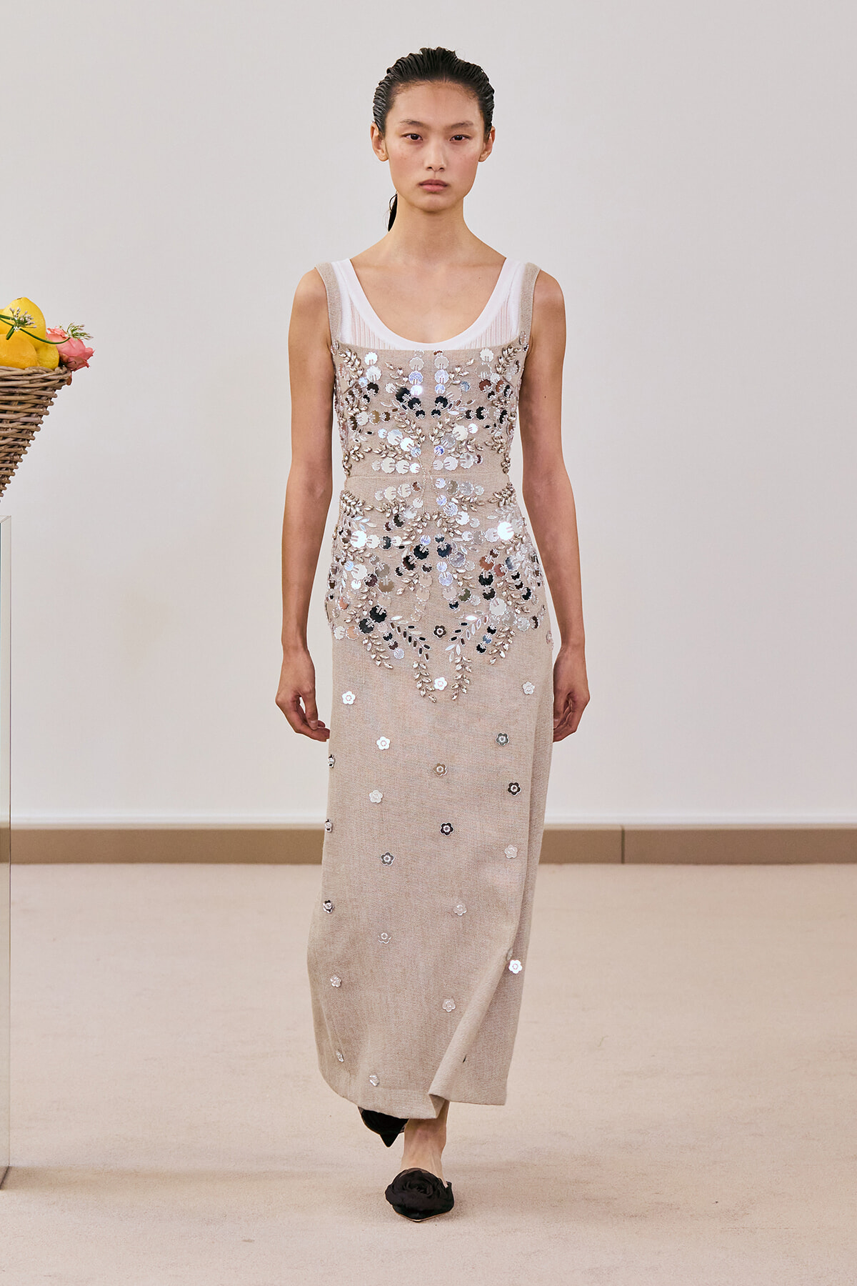 Full-length beige sleeveless gown with silver sequin floral embroidery, worn over a white mesh top, model walking on a runway.