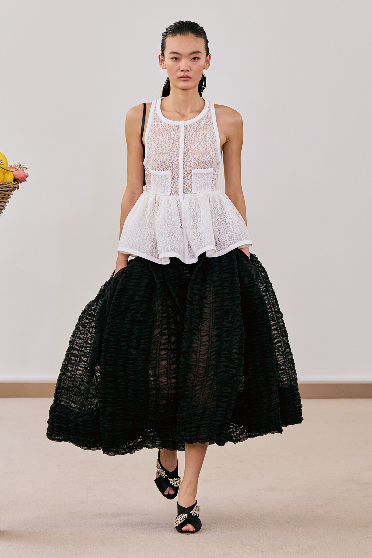 Model wearing a white lace sleeveless peplum top with a voluminous black textured midi skirt, walking forward in a fashion show setting.