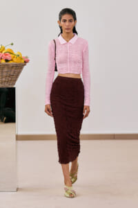 Model wearing a pink ruched cropped sweater with a collared neckline and a burgundy ruched midi skirt, standing in a neutral studio with a fruit basket nearby.
