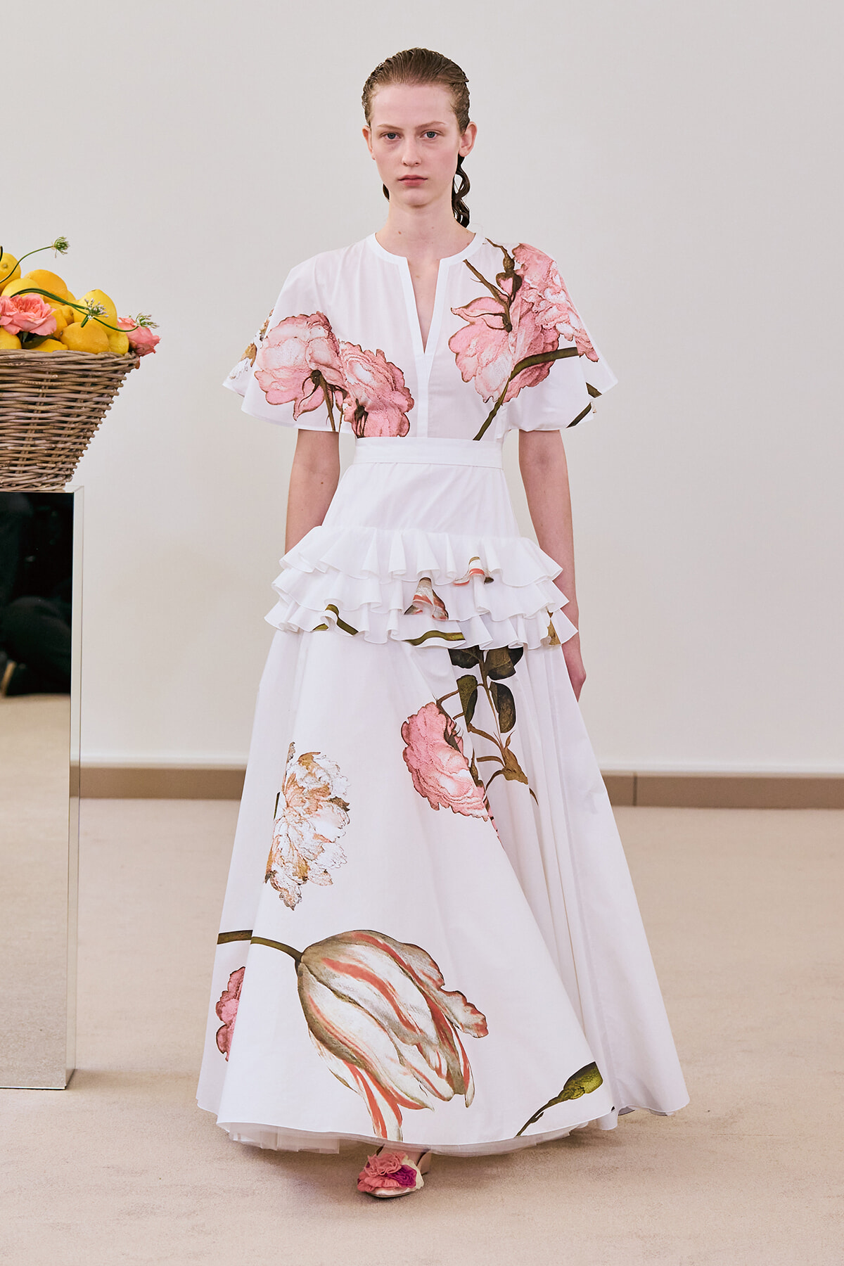 Model wearing a white floral gown with pink blossoms and layered ruffles, standing indoors beside a basket of lemons.