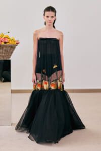 Model wearing a strapless black mesh gown with a fruit-print hem, standing in a minimalist room with a fruit basket beside her.