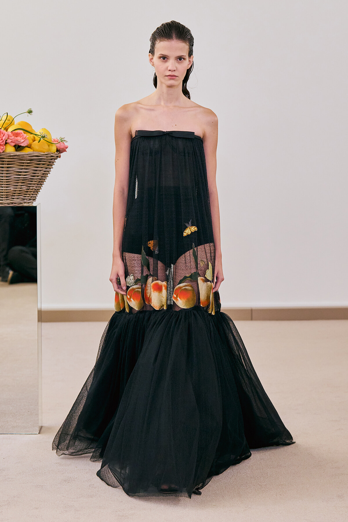Model wearing a strapless black mesh gown with a fruit-print hem, standing in a minimalist room with a fruit basket beside her.