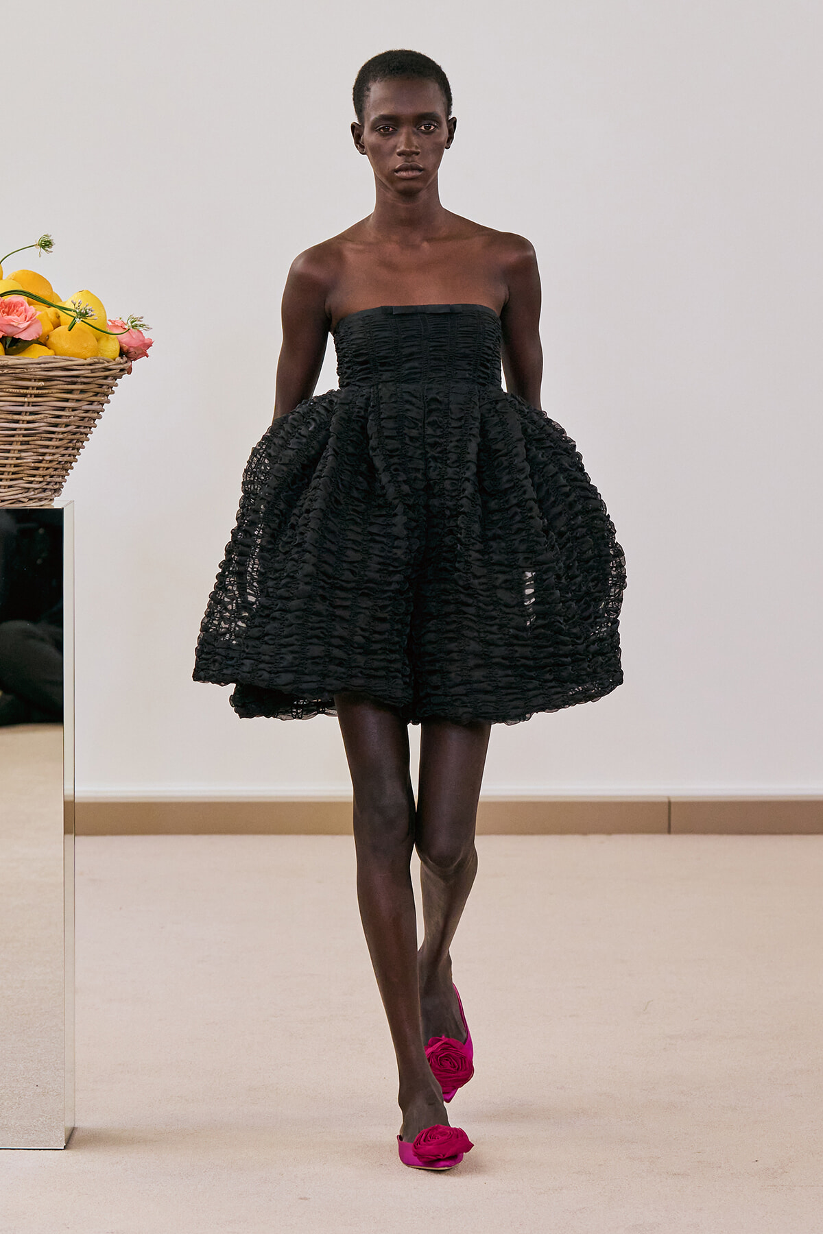 Model wearing a strapless black textured mini dress with a full, voluminous skirt on a runway; pink flower-embellished heels visible.