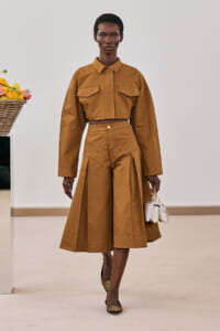 Model in a camel cropped shirt and high-waisted wide-leg skirt with pleats, carrying a small white handbag.