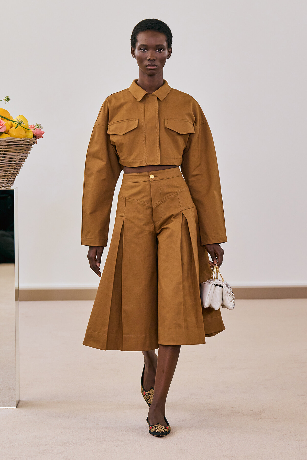 Model in a camel cropped shirt and high-waisted wide-leg skirt with pleats, carrying a small white handbag.