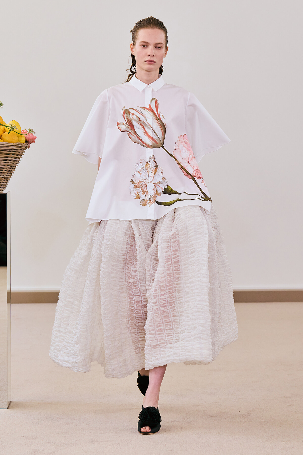 Model wearing a white oversized blouse with large pink flower print paired with a voluminous textured pink skirt and black open-toe sandals.