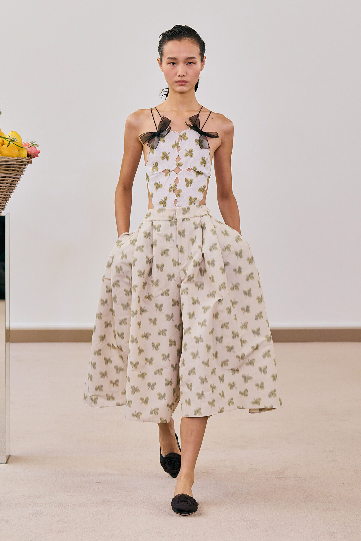 Model walking the runway in a cream midi dress with a green butterfly print and a cutout bodice with black bow straps; paired with black flower-embellished flats.