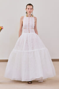 Model wearing a full-length white lace gown with a tiered skirt and scalloped hem, accessorized with large gold earrings.