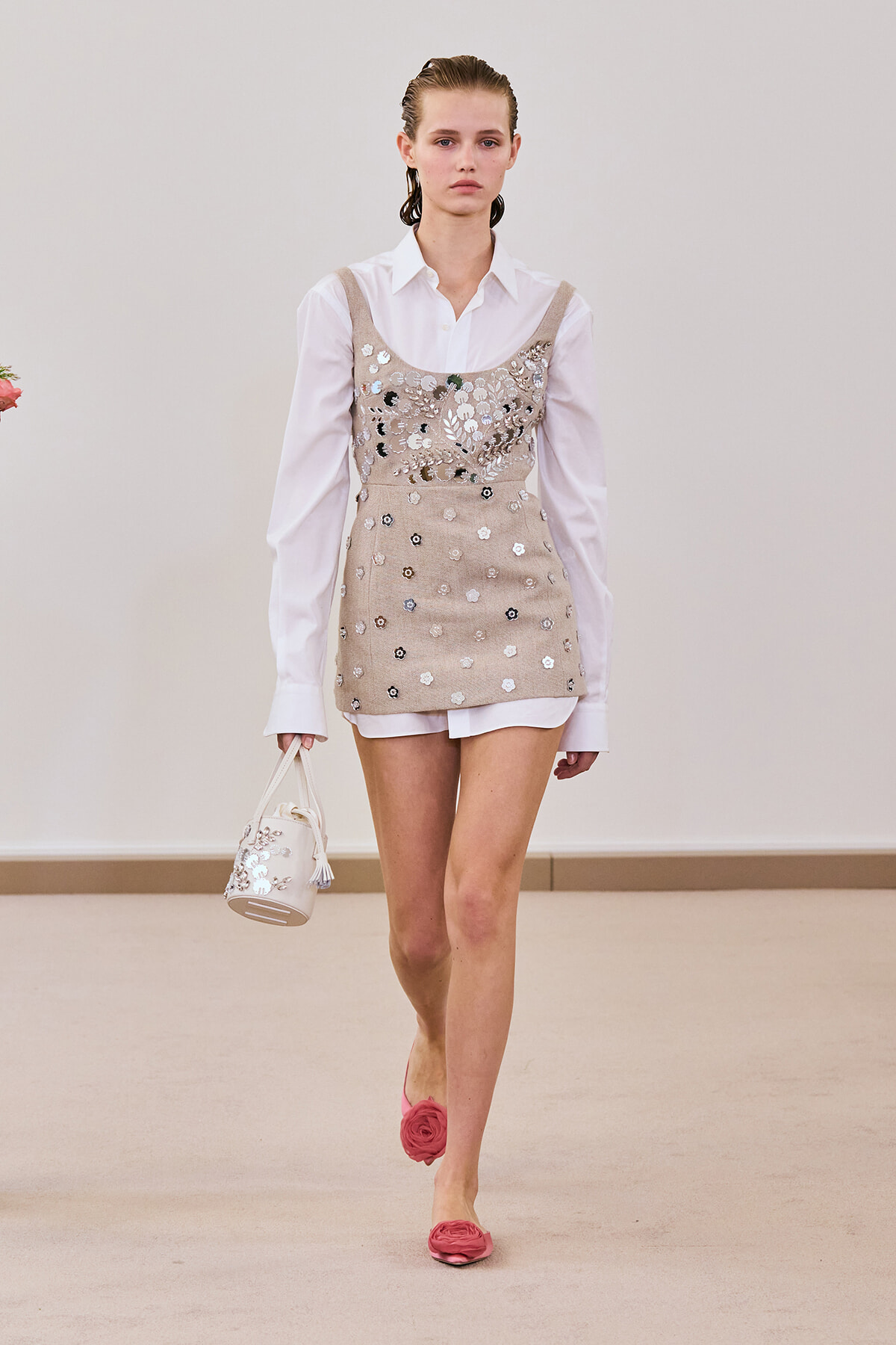 Model walking in a beige sequin slip worn over a white button-down shirt, holding a small white handbag and wearing pink floral flats.