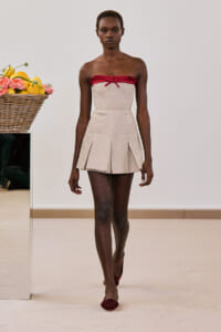 Model on a fashion runway wearing a beige strapless dress with a red ruffle neckline and pleated skirt, burgundy flats; a wicker basket of lemons sits to the left.