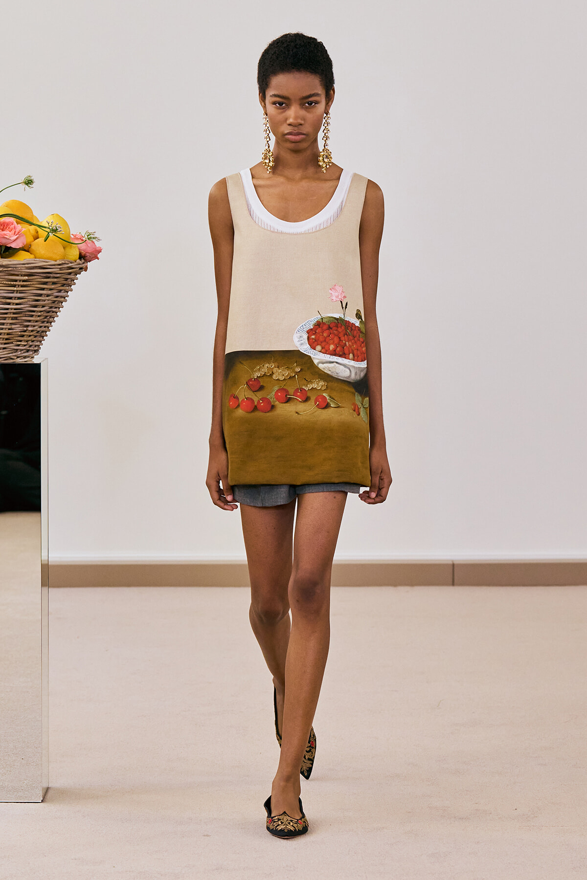 Model walking a runway in a sleeveless beige dress with a two-tone hem showing a fruit still-life print; gold chandelier earrings.
