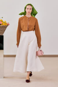 Model walking in a studio wearing a brown button-front blouse, white wide-leg pants, a green headscarf, burgundy flats, and a pink handbag.