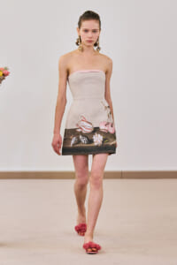 Model walking in a strapless beige mini dress with a dark floral hem, pink trim at top, gold statement earrings, and pink flower sandals.