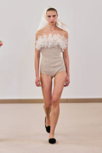 Model walking the runway in a beige off-the-shoulder bodysuit with a fluffy white tulle neckline and a draped white headscarf, black flats.