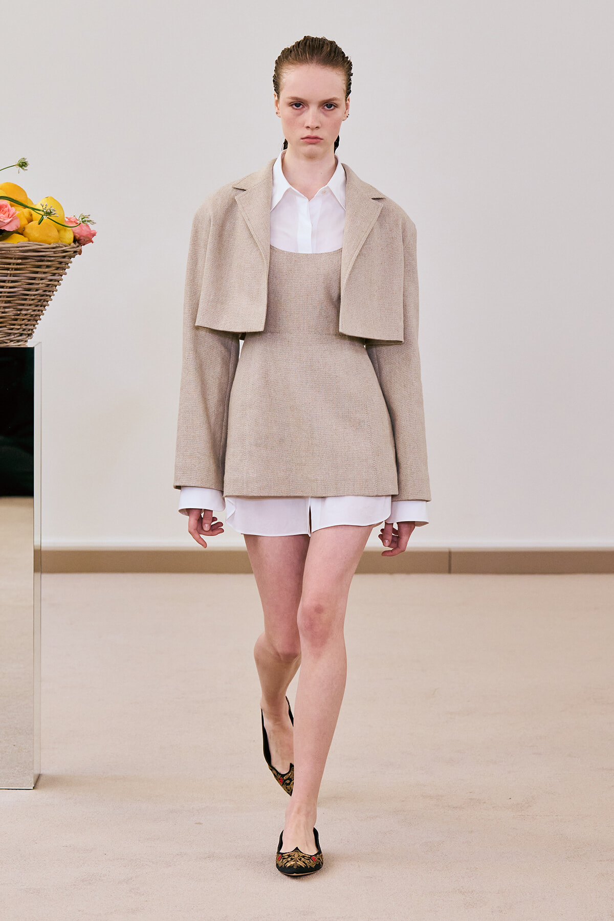 Model walking down a runway in a beige layered outfit: cropped cape over a white shirt and matching tunic-like dress, white shirt cuffs peeking out, wearing embroidered flats; a vase of flowers sits to the left.