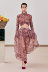 Model wearing a pink floral textured two-piece outfit (crop top and long skirt) walking on a runway with a basket of lemons nearby left.