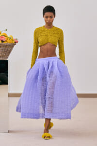 Model walking a runway in a mustard yellow ruched cropped top and lavender voluminous skirt; lemons in a basket visible at left.