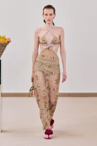 Model walking a fashion runway in a sheer beige lace dress with pink floral embroidery and crisscross straps, carrying a small pink purse and wearing pink flower-adorned sandals.