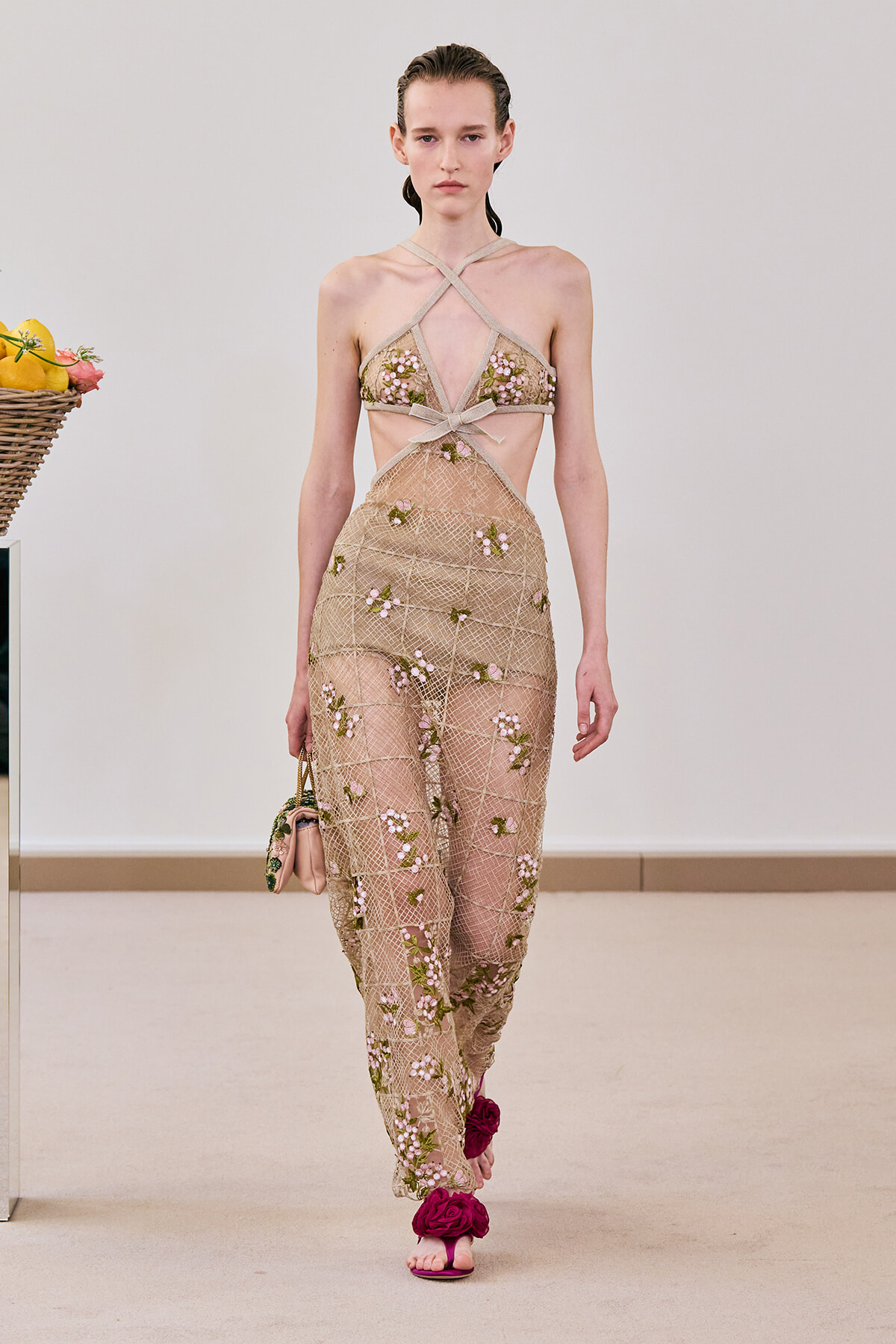 Model walking a fashion runway in a sheer beige lace dress with pink floral embroidery and crisscross straps, carrying a small pink purse and wearing pink flower-adorned sandals.