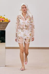 Model walks a fashion show wearing a sheer floral-embroidered pink coat over white undergarments, paired with floral flats and a white headscarf; basket of lemons visible nearby.