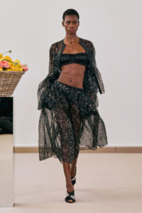 Model walks the runway in a sheer black floral set with a ruffled black bandeau top and matching voluminous skirt, mid-stride, on a light background.