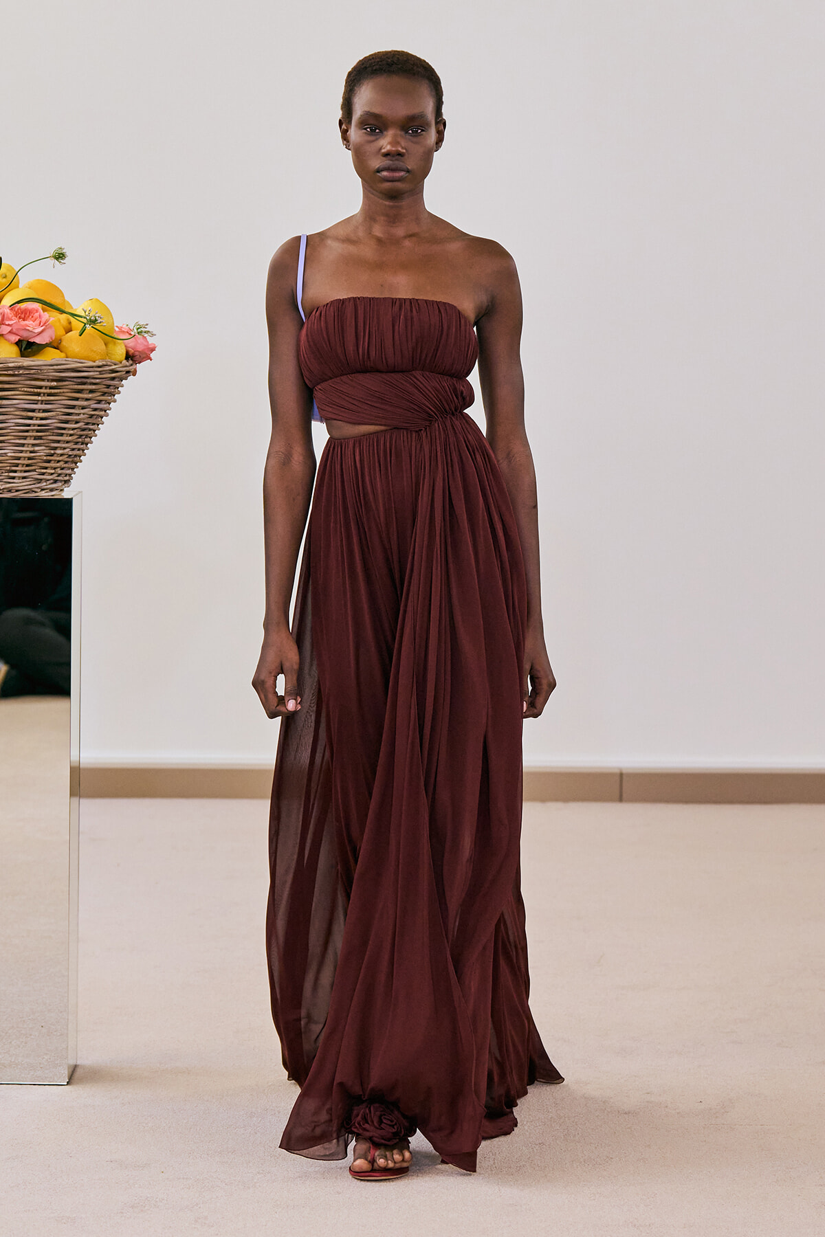Model wearing a burgundy draped gown with one strap, standing on a runway beside a wicker basket of lemons and flowers.