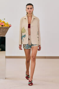 Model walking a runway in a beige blazer with tropical embroidery, matching shorts, burgundy sandals, and bold gold earrings.