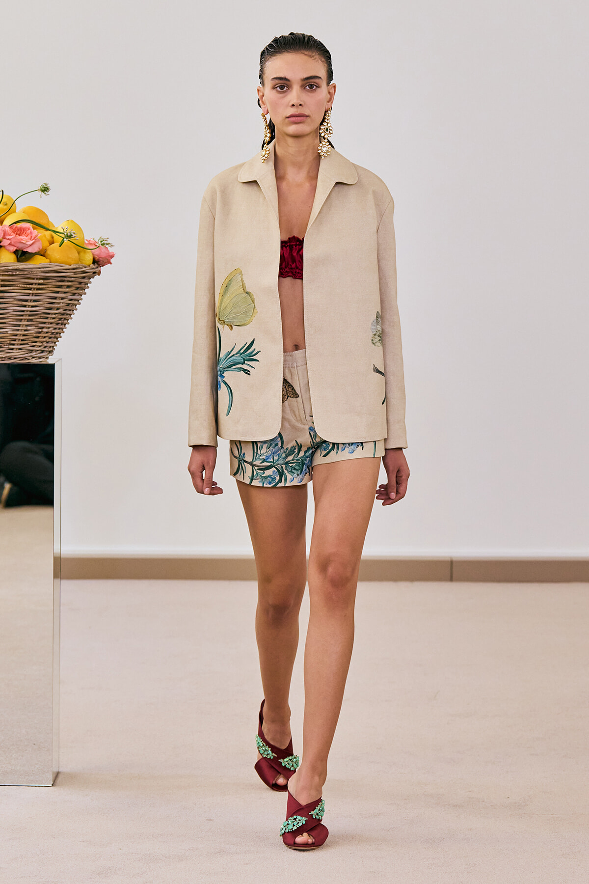 Model walking a runway in a beige blazer with tropical embroidery, matching shorts, burgundy sandals, and bold gold earrings.