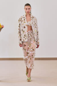 Model walking in a beige floral suit: matching jacket and wide-leg cropped pants with a ruffled beige top, accessorized with large gold earrings and green embellished sandals.