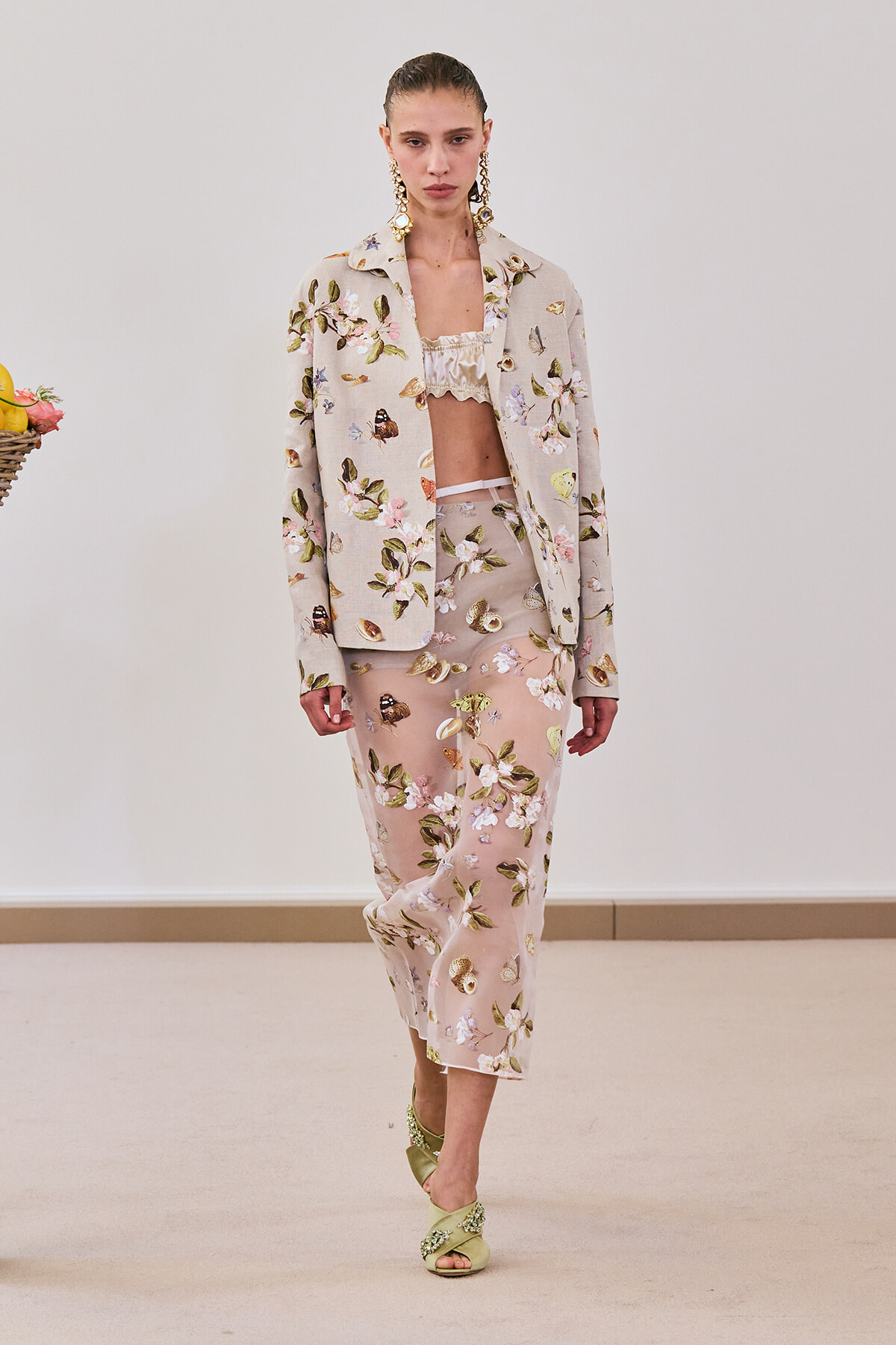 Model walking in a beige floral suit: matching jacket and wide-leg cropped pants with a ruffled beige top, accessorized with large gold earrings and green embellished sandals.