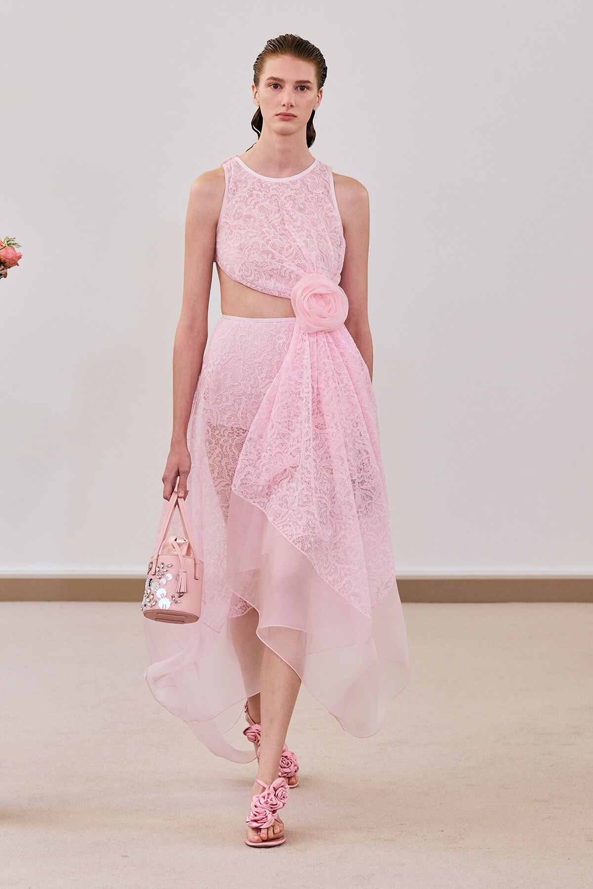 Runway model wearing a pink lace two-piece dress with a large fabric rose belt, carrying a pink embellished handbag and pink sandals.
