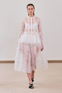 Model wearing a sheer white lace dress with floral patterns, peplum waist, and black jeweled sandals on a runway show.