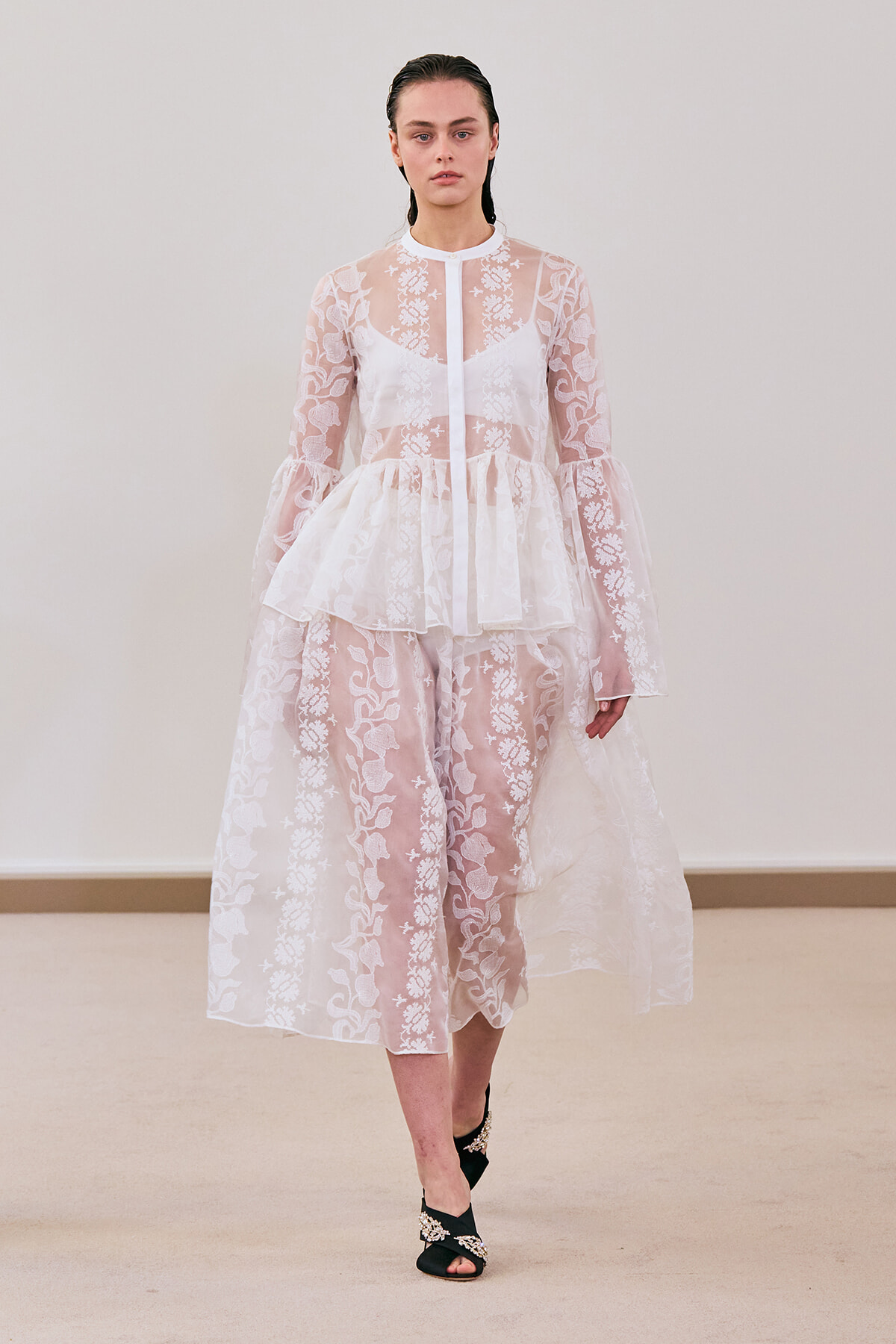 Model wearing a sheer white lace dress with floral patterns, peplum waist, and black jeweled sandals on a runway show.