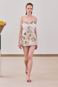 Model walking a runway in a beige floral mini dress with a large white bow at the bust and pale pink floral flats.