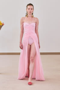 Runway model in a light pink, ruched gown with a high front slit and thin straps, wearing large gold earrings.