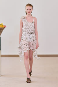 Model wearing a pale pink floral wrap dress with a one-shoulder drape and high-low ruffled hem, walking on a runway with neutral backdrop.