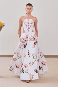 Model in a white, floor-length floral gown with pink blossoms and thin straps on a runway, with a fruit basket visible to the left.