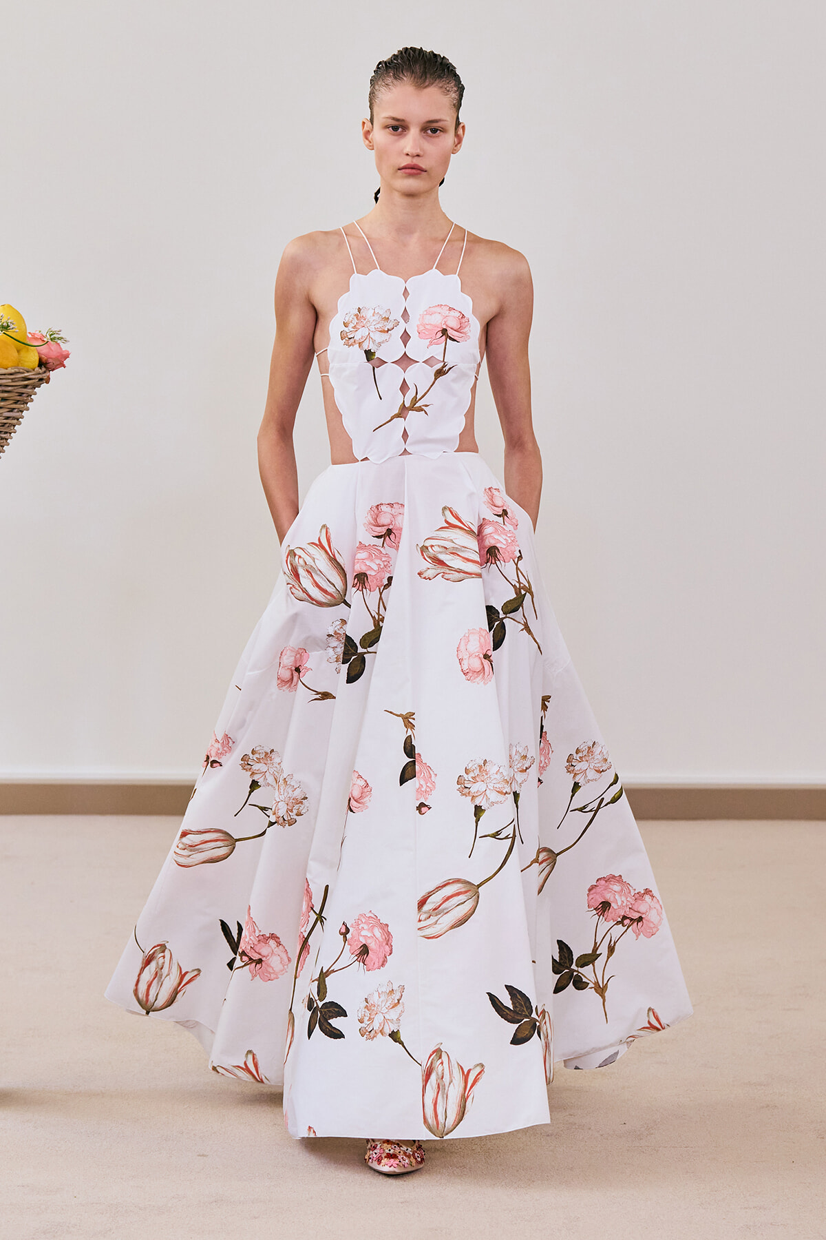 Model in a white, floor-length floral gown with pink blossoms and thin straps on a runway, with a fruit basket visible to the left.